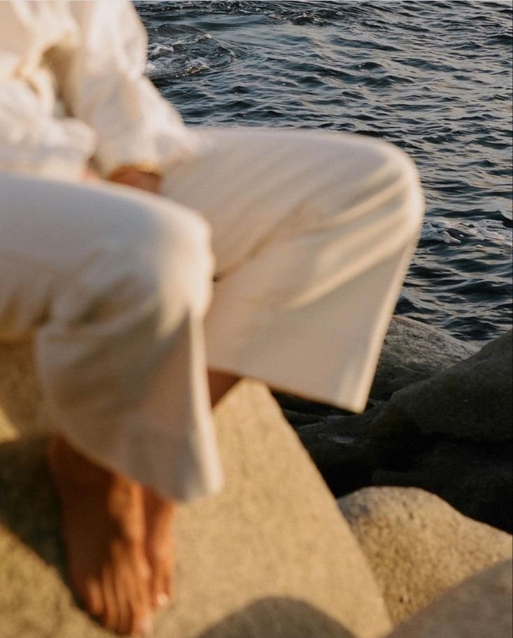 A photograph of a woman's legs as she sits on a pile of rocks near the sea shore.